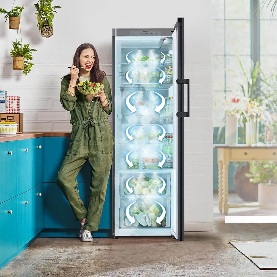 Równomierne chłodzenie w lodówce Samsung z oferty sprzedażowej Media Expert zapewnia system chłodzący All-Around Cooling Kobieta w zielonym kombinezonie stoi obok otwartej lodówki, jedząc sałatkę. Wnętrze lodówki jest wypełnione różnymi produktami spożywczymi, a na drzwiach widoczne jest logo \'SAMSUNG\'.