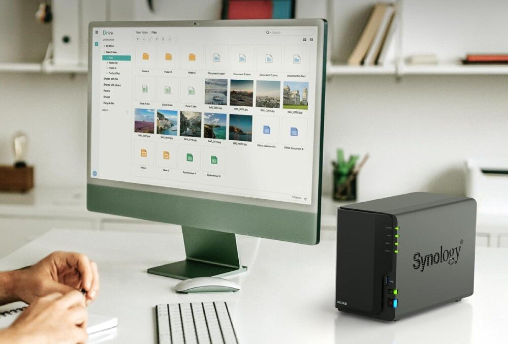 Serwer plików SYNOLOGY DiskStation DS224+ Zdjęcie przedstawia biurko, na którym znajduje się monitor z otwartą aplikacją do zarządzania plikami. Obok monitora stoi niewielki serwer plików, opis