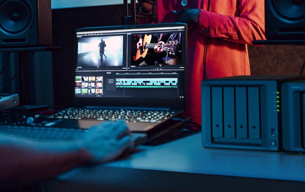 Serwer plików SYNOLOGY DS923+ Osoba pracuje przy komputerze z otwartym oprogramowaniem do edycji wideo. Na ekranie widać dwa podglądy filmów i oś czasu edycji. Obok laptopa stoi serwer plików z kilkoma zatokami na dyski, opis