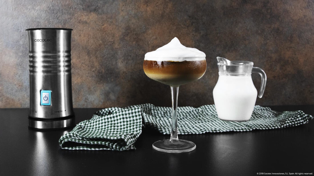 Na obrazie znajduje się metalowy spieniacz do mleka z widocznym przyciskiem zasilania oraz szklanka z napojem z pianką mleczną. Obok leży zielona ściereczka i stoi dzbanek z mlekiem.