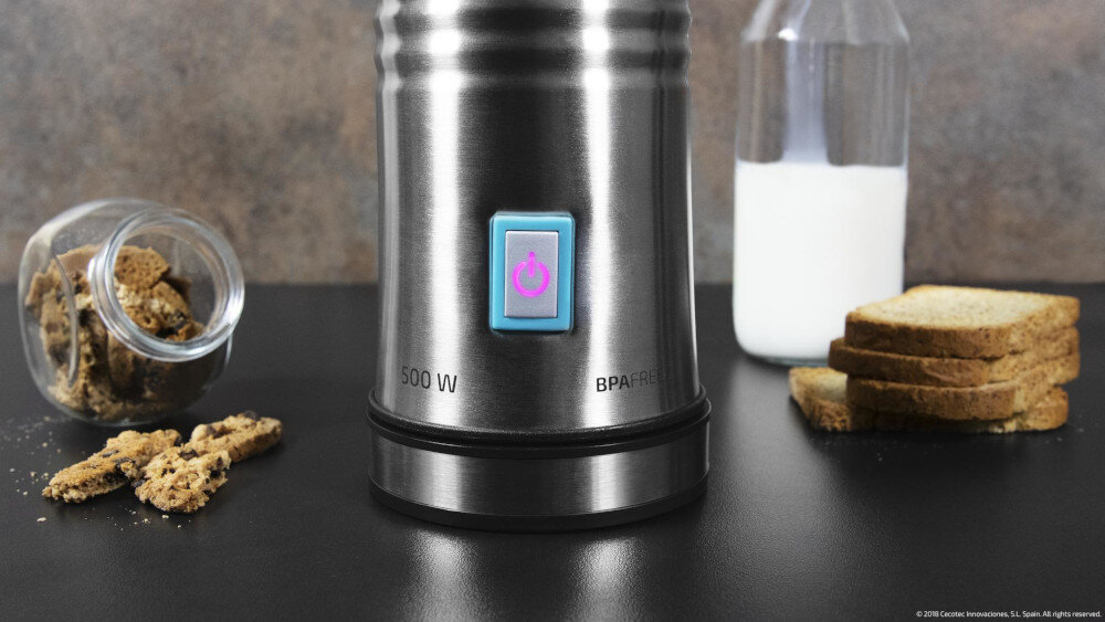 Na obrazie widoczny jest metalowy spieniacz z niebieskim przyciskiem i napisami \'500 W\' oraz \'BPA FREE\'. Obok znajdują się słoik z pokruszonymi ciastkami, butelka mleka i stos kromek chleba.