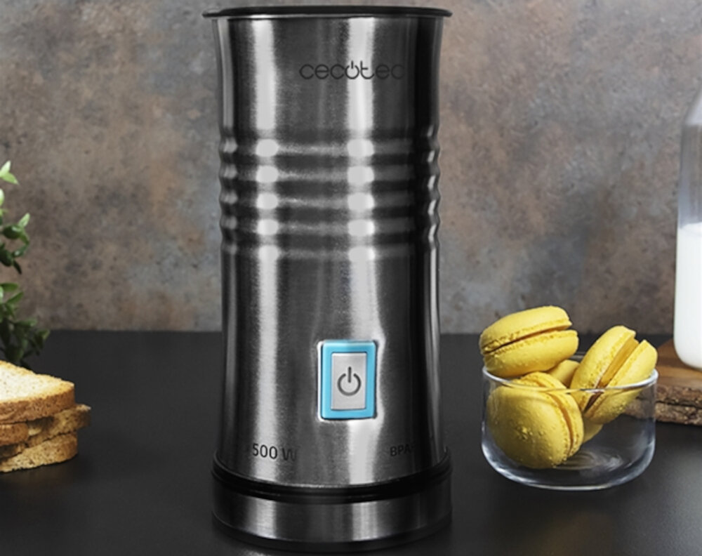 Na obrazie widoczny jest metalowy spieniacz do mleka z logo \'cecotec\' i przyciskiem zasilania. Obok znajdują się żółte makaroniki w szklanej miseczce oraz kromki chleba.