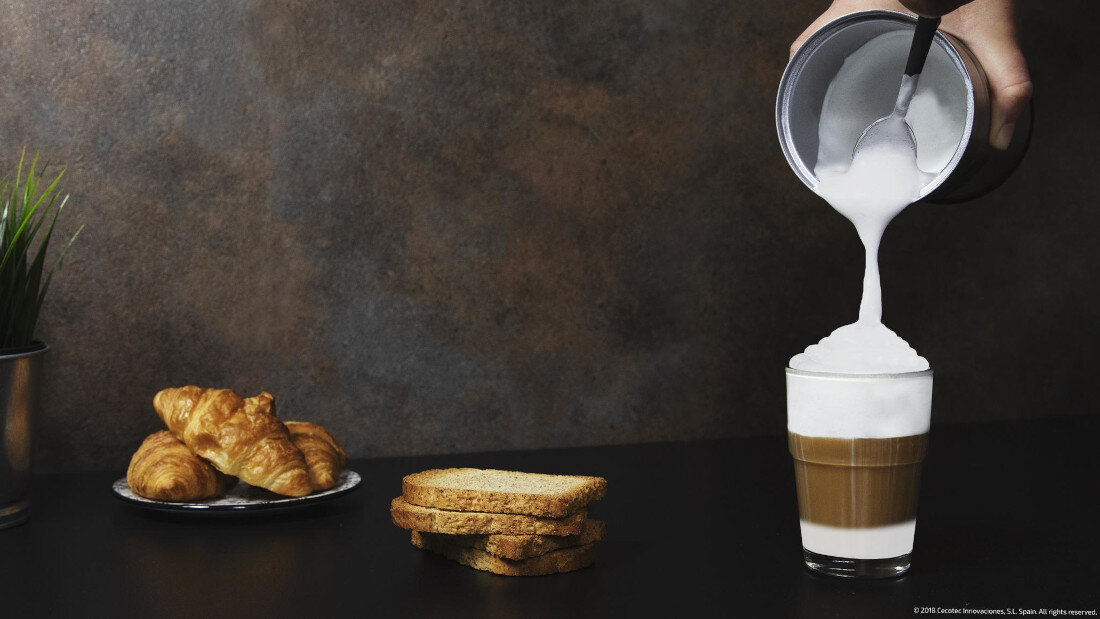 Na stole znajduje się szklanka z kawą, do której nalewana jest spieniona mleczna pianka z metalowego naczynia. Obok leży talerz z croissantami i kilka kromek chleba, a w tle widać doniczkę z rośliną.