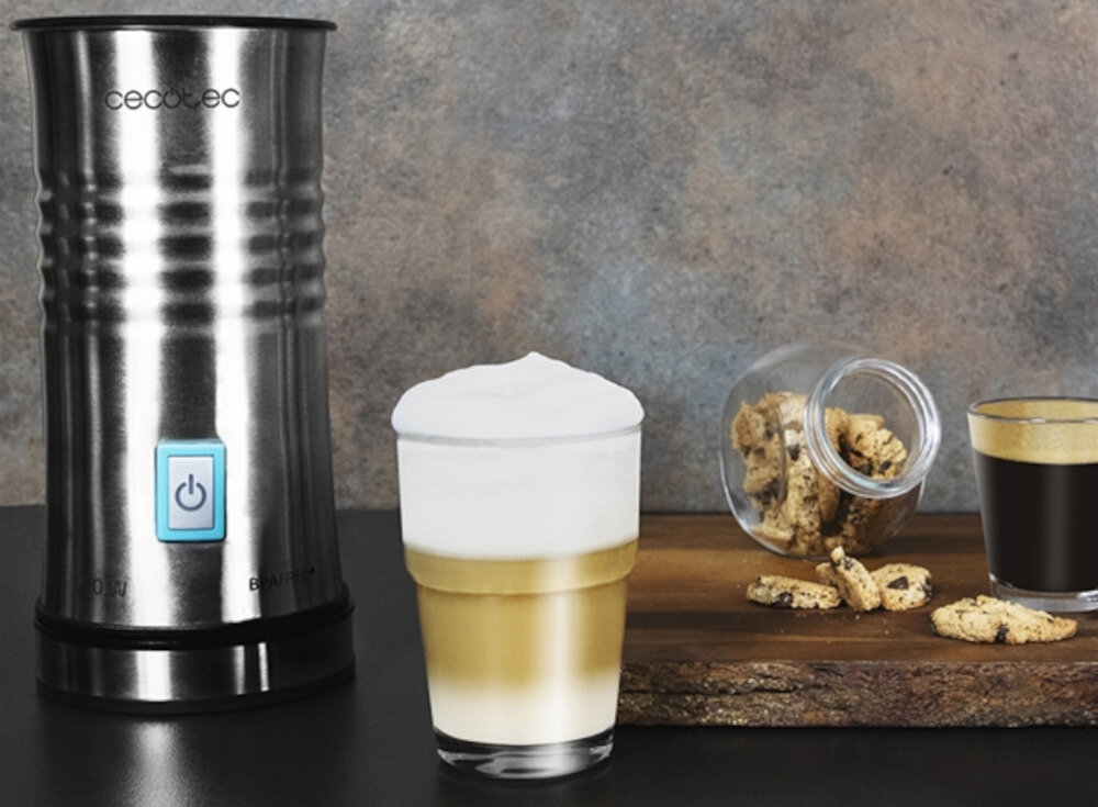 Na obrazie widoczny jest metalowy spieniacz do mleka z logo \'cecotec\' oraz szklanka z warstwowym napojem mlecznym. Obok znajduje się deska z rozsypanymi ciasteczkami i szklanką czarnej kawy.