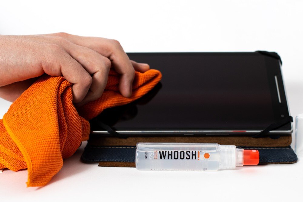 Osoba czyści ekran tabletu pomarańczową ściereczką. Przed tabletem leży butelka z napisem \'WHOOSH!\'.