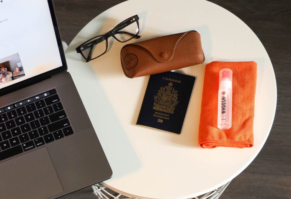 Na okrągłym stole znajduje się laptop, okulary w etui, paszport z napisem \'CANADA PASSPORT\', oraz pomarańczowa ściereczka z buteleczką WHOOSH!.