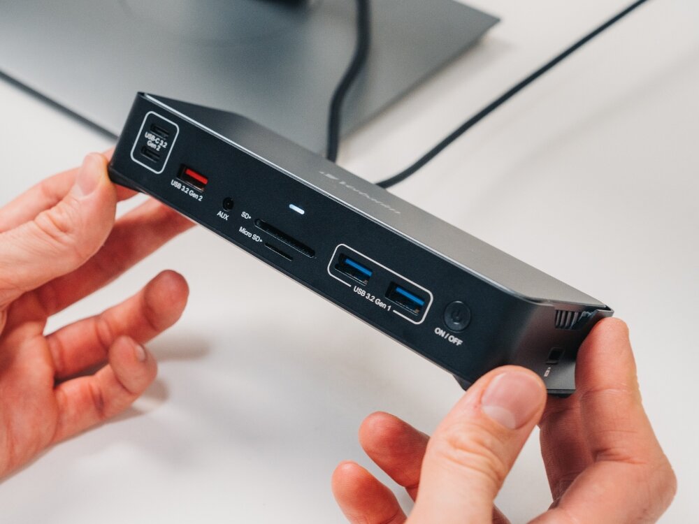 Osoba trzyma stację dokującą z różnymi portami, w tym USB 3.0, USB-C, slot na karty SD i microSD oraz przycisk ON/OFF. Na urządzeniu widoczne są napisy: \'USB 3.0 Gen 2\', \'USB 3.0 Gen 1\', \'ON/OFF\'.