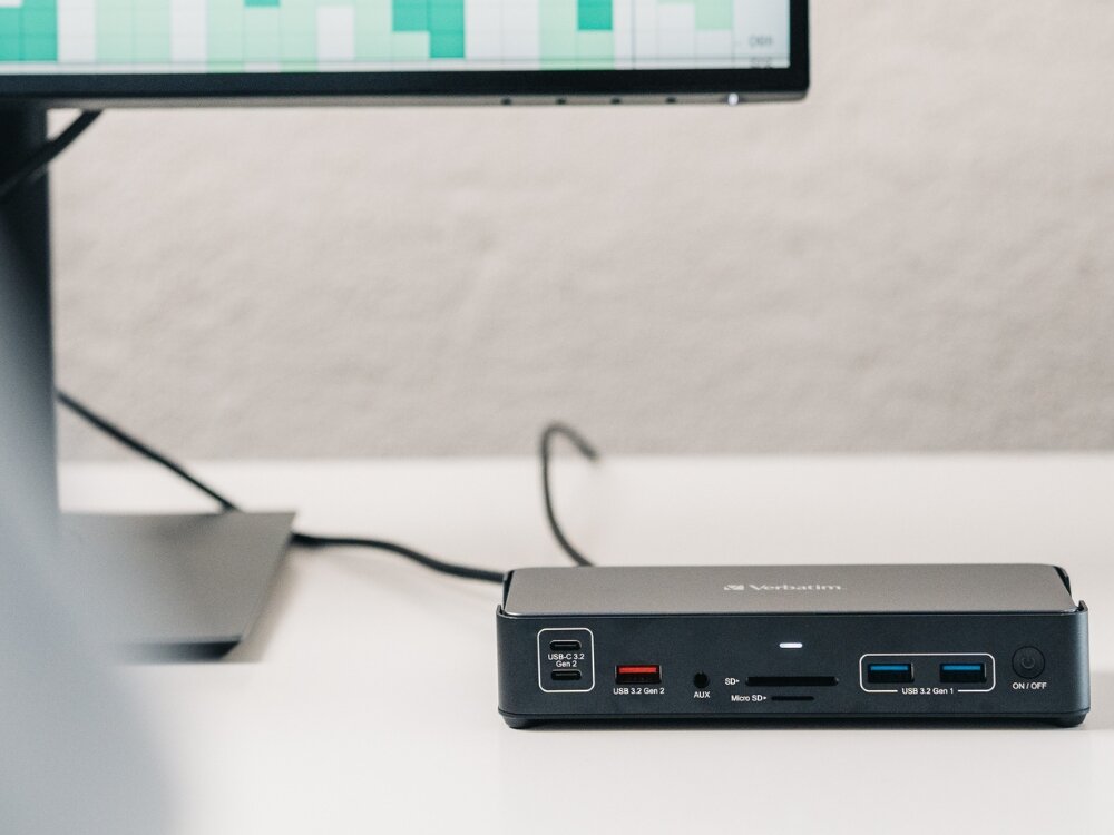 Na biurku znajduje się stacja dokująca z logo Verbatim, posiadająca różne porty, w tym USB-C, USB 3.2 Gen 1, SD i Micro SD. W tle widoczny jest fragment monitora z wykresem słupkowym.