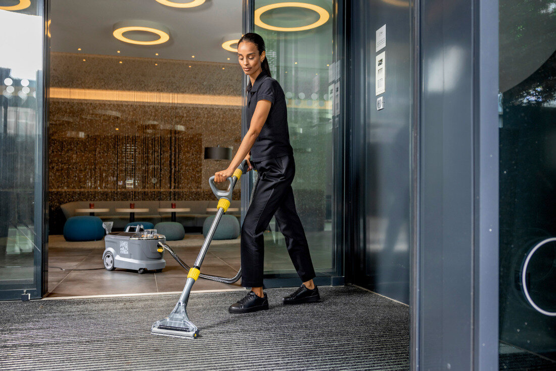 Osoba używa odkurzacza ekstrakcyjnego na dywanie przy wejściu do nowoczesnego budynku. W tle widoczny jest odkurzacz z logo KARCHER.