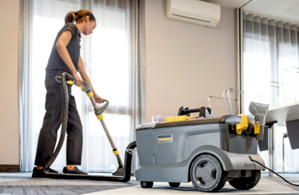 Osoba używa odkurzacza KARCHER Professional Puzzi 10/1 w jasnym pomieszczeniu z dużymi oknami. Na odkurzaczu widoczne jest logo KARCHER.