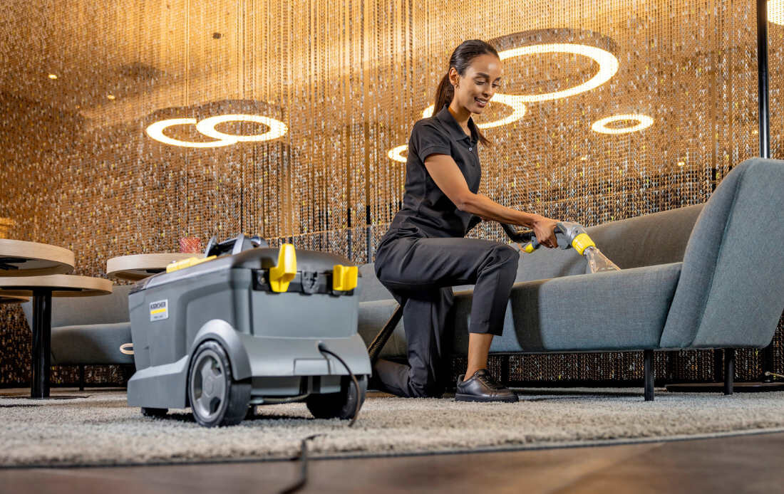 Osoba używa odkurzacza do czyszczenia sofy w eleganckim wnętrzu z nowoczesnym oświetleniem. Na odkurzaczu widoczne jest logo KARCHER.