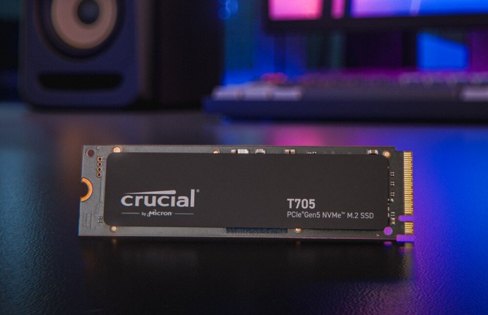 Dysk CRUCIAL T705 Dysk SSD oznaczony jako 'T705 PCIe Gen5 NVMe M.2' stojący pionowo na ciemnej powierzchni, w tle rozmyty monitor i głośnik z kolorowym oświetleniem, opis