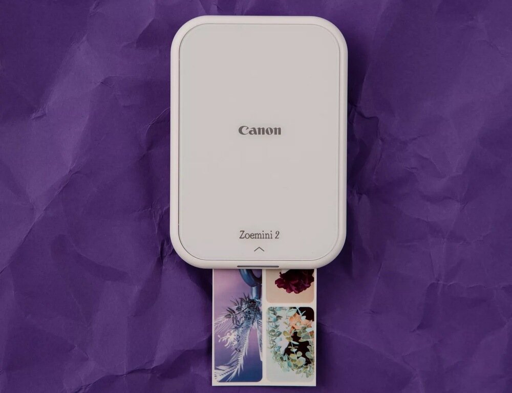 Na tle fioletowego materiału znajduje się drukarka z widocznym logo \'Canon\' oraz napisem \'Zoemini 2\'. Z urządzenia wysuwa się kolorowe zdjęcie przedstawiające kwiaty.