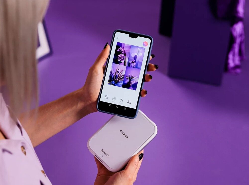 Osoba trzyma smartfon z wyświetlonymi zdjęciami kwiatów oraz różowo-białą drukarkę Canon Zoemini 2. Tło jest fioletowe, a na drukarce widoczne jest logo \'Canon\'.