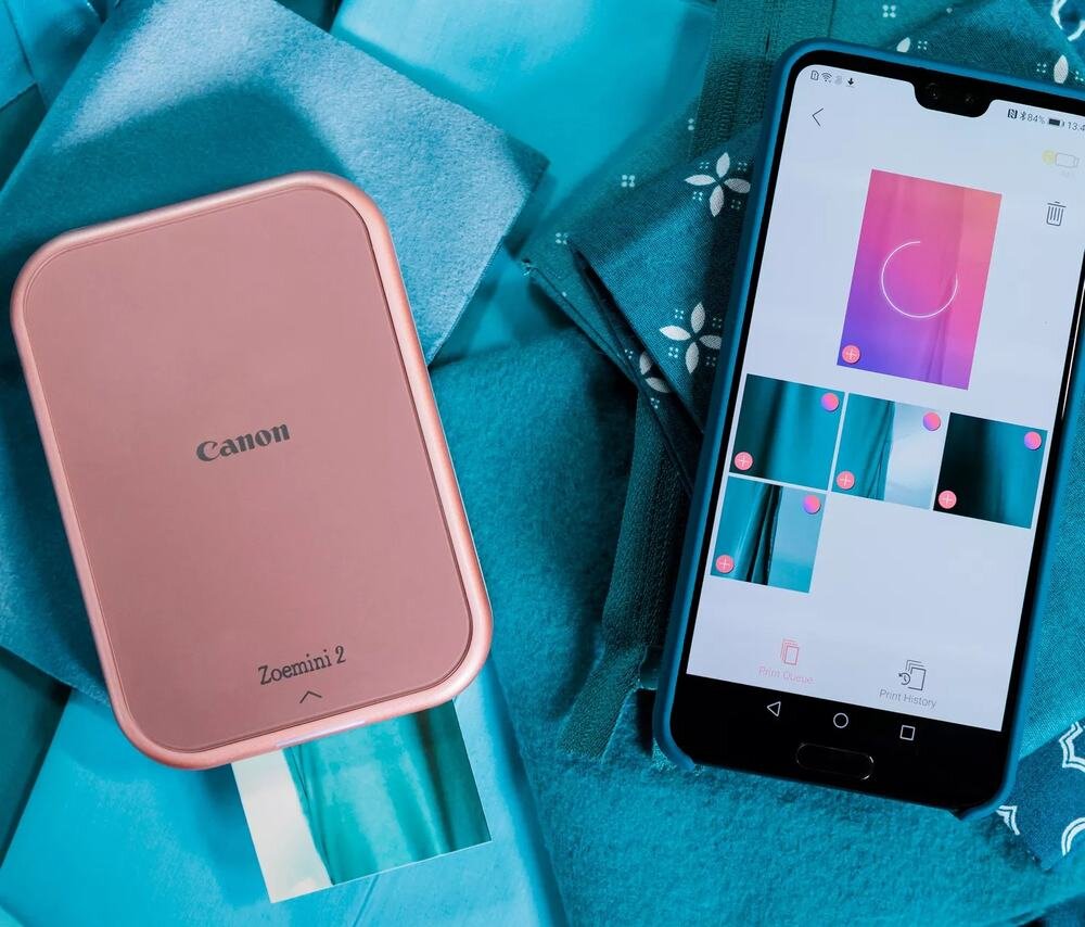 Na obrazie widoczna jest różowo-biała drukarka z napisem \'Canon Zoemini 2\' oraz smartfon z aplikacją do drukowania zdjęć. Tło jest w odcieniach niebieskiego z delikatnym wzorem.