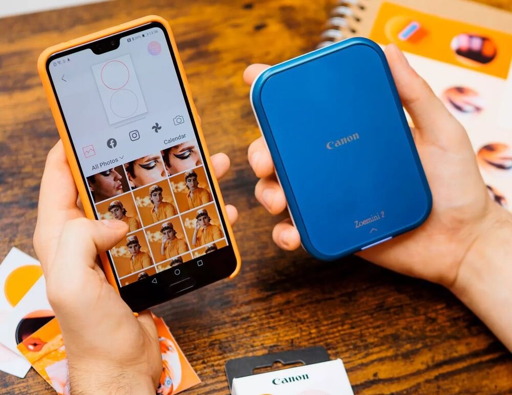 Osoba trzyma smartfon z aplikacją do edycji zdjęć oraz niebieskie urządzenie z napisem \'Canon Zoemini 2\'. Na stole leży opakowanie z logo \'Canon\'.