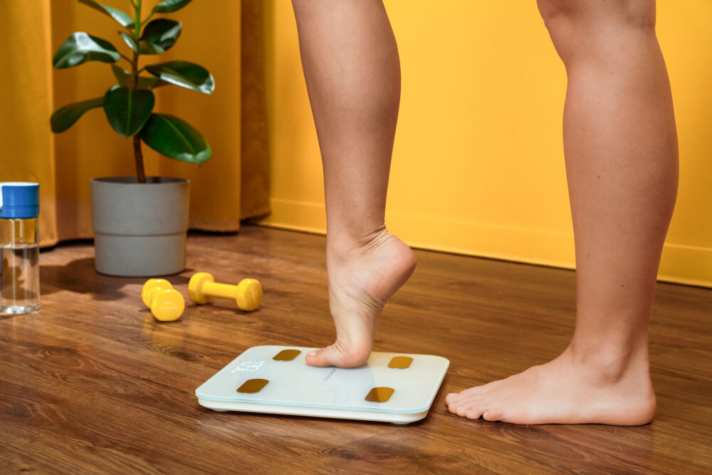 Waga łazienkowa GÖTZE & JENSEN Analizator BT BS700W Biały osoba wchodząca na wagę w tle widoczna butelka na wodę i hantelki