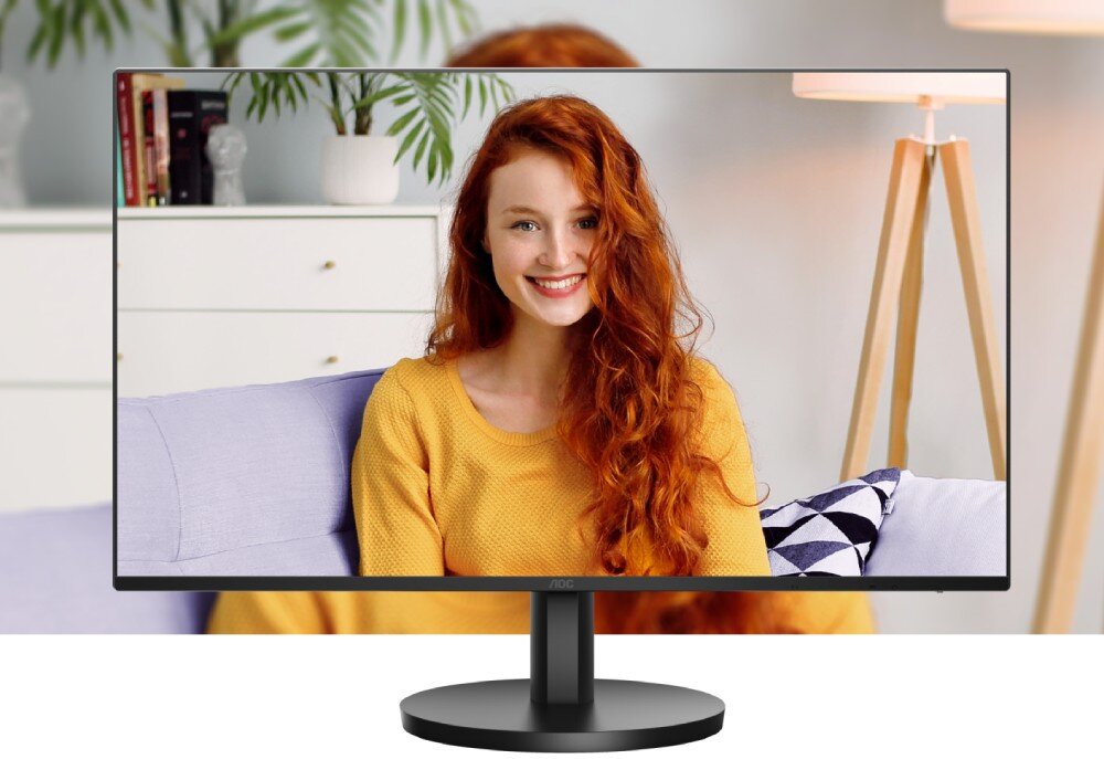 Monitor AOC 27B3HA2 Monitor na biurku z wyświetlonym obrazem kobiety o rudych włosach w żółtym swetrze, siedzącej na kanapie. W tle widoczne są rośliny, lampa i biała komoda, opis