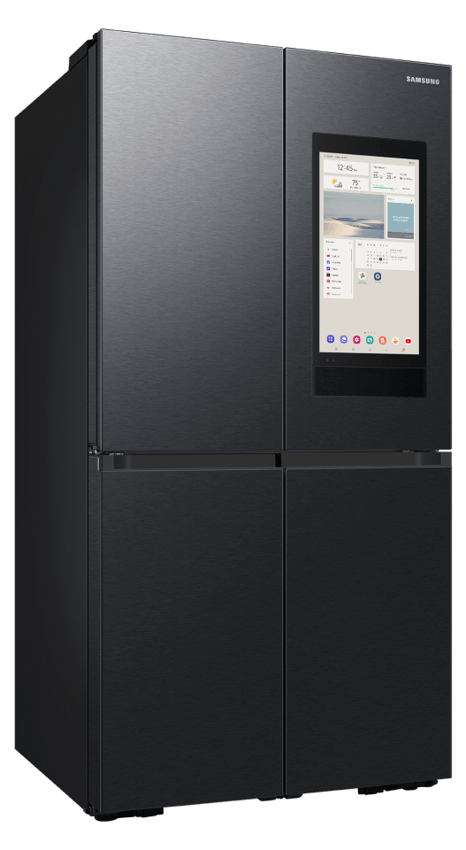 Lodówka w kolorze grafitowej stali z czterema drzwiami i wbudowanym ekranem dotykowym na jednym z górnych paneli. Na ekranie widoczne są różne aplikacje i informacje, a w prawym górnym rogu znajduje się logo \'SAMSUNG\'.