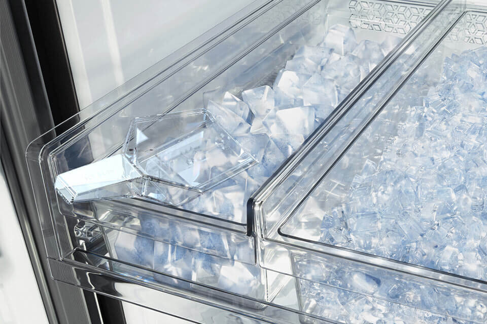 Obraz przedstawia wnętrze lodówki z przezroczystymi pojemnikami wypełnionymi kostkami lodu. W jednym z pojemników znajduje się plastikowa łopatka do lodu.