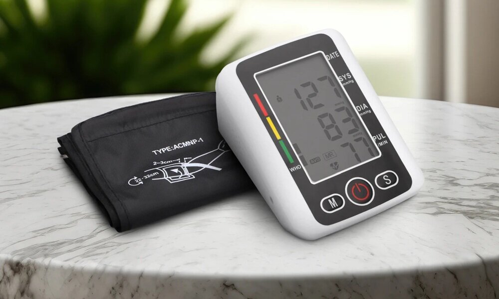 Na marmurowym stole znajduje się ciśnieniomierz z wyświetlaczem pokazującym wartości 127, 83 i 73 oraz mankiet z napisem \'TYPE: ACMNP-A\'.