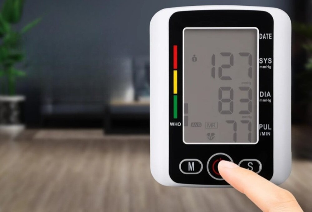 Na obrazie widoczny jest ciśnieniomierz z wyświetlaczem pokazującym wartości: 127 SYS, 83 DIA, 77 PUL. Obok wyświetlacza znajdują się kolorowe wskaźniki oraz przyciski oznaczone literami M i S.