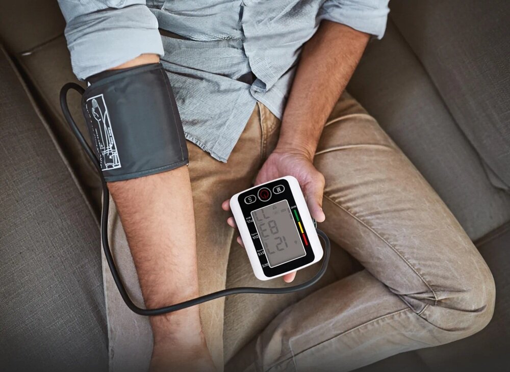 Osoba siedzi na kanapie z ciśnieniomierzem na ramieniu, trzymając w ręku urządzenie z wyświetlaczem pokazującym cyfry. Na mankiecie widoczny jest schemat zakładania.