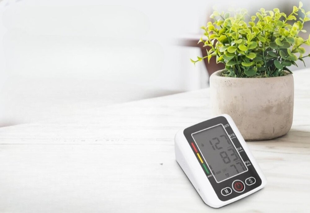 Na białym stole stoi ciśnieniomierz z wyświetlaczem pokazującym wartości 127, 83 i 77. Obok znajduje się doniczka z zieloną rośliną.