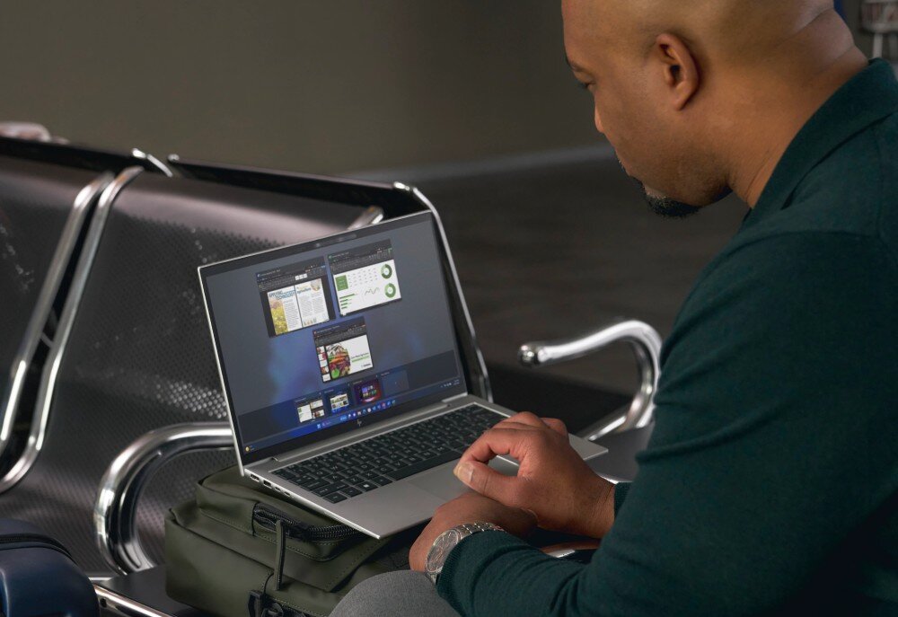 Laptop HP EliteBook 865 G10 Mężczyzna siedzi na lotnisku na metalowej ławce i pracuje na laptopie, który spoczywa na jego torbie. Na ekranie widoczne są otwarte okna aplikacji, opis
