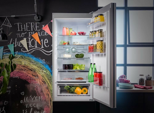 Otwarta lodówka z różnorodnymi produktami spożywczymi na półkach, w tym warzywa, napoje i słoiki. W tle widoczna jest kolorowa tablica z napisem \'THERE IS NO PLACE like HOME\'.