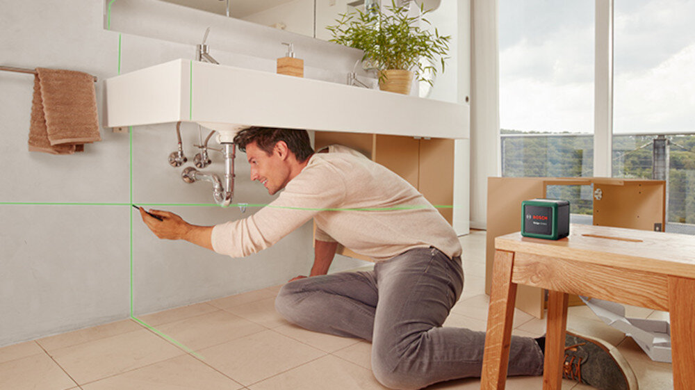 Laser krzyżowy BOSCH Quigo Green Set 0603663CZ1 pomieszczenie łazienka osoba wyznacza linie na ścianie w tle okno obok stolik na stoliku laser do prac remontowych montażowych i dekoracyjnych zastosowanie w domowych obowiązkach i w profesjonalnym wykończeniu wnętrz estetyczność oszczędność czasu