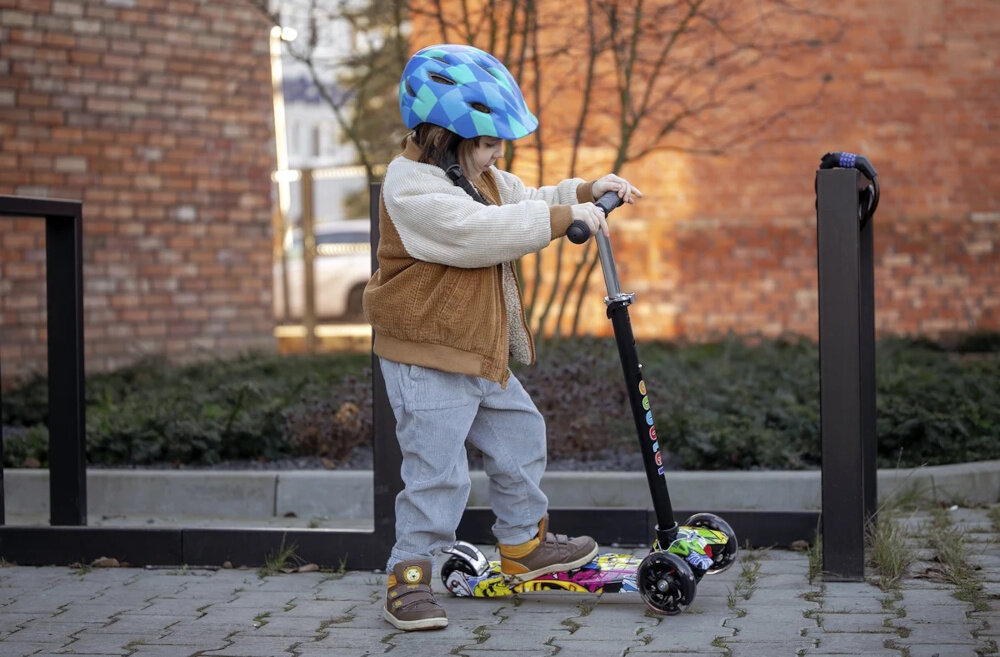 Hulajnoga dla dzieci EXTRALINK Baby Adventure droga chodnik dziecko opiera stopę o platformę hulajnogi w tle budynki z cegły zabawa rozwój ergonomia bezpieczeństwo