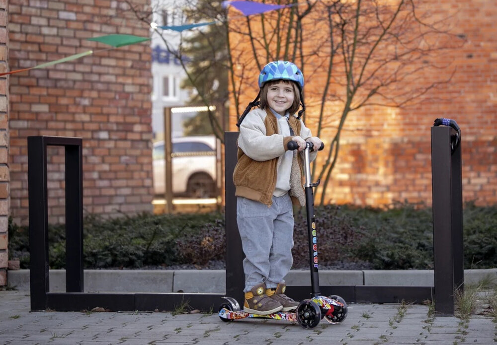 Hulajnoga dla dzieci EXTRALINK Baby Adventure droga chodnik dziecko opiera stopę o platformę hulajnogi w tle budynki z cegły lekka platforma wykonana z wysokiej jakości tworzywa sztucznego antypoślizgowa powierzchnia pewne oparcie dla stóp szeroki podest obciążenie do 50 kg