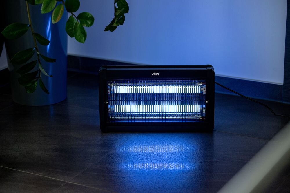 Na podłodze stoi lampa owadobójcza emitująca niebieskie światło. Na urządzeniu widoczne jest logo \'VAYOX\'.