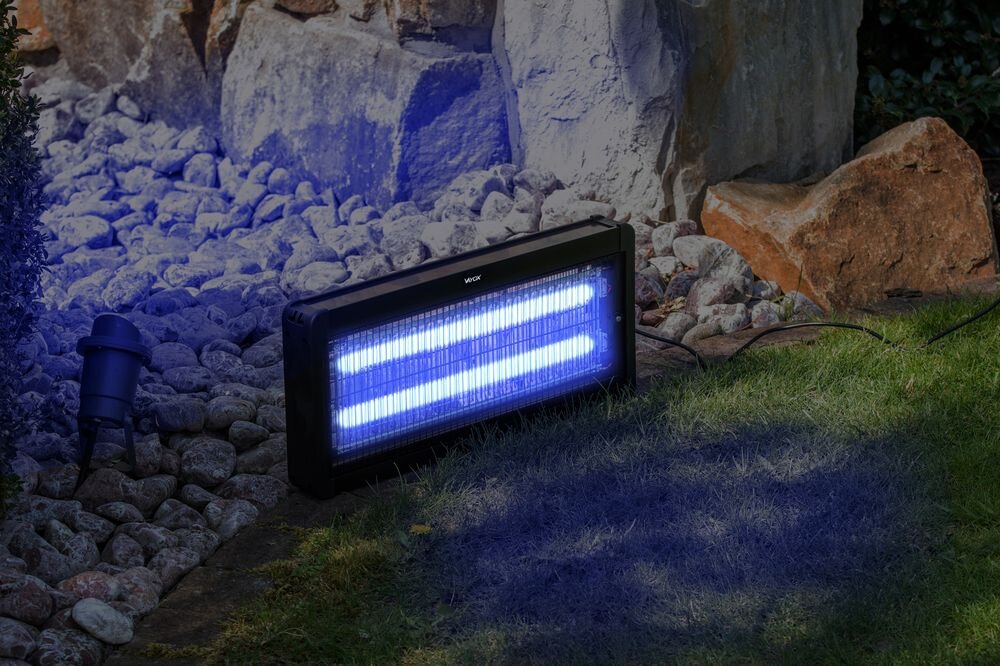 Na trawie i kamieniach stoi lampa owadobójcza emitująca niebieskie światło. Na obudowie widoczne jest logo \'VAYOX\'.