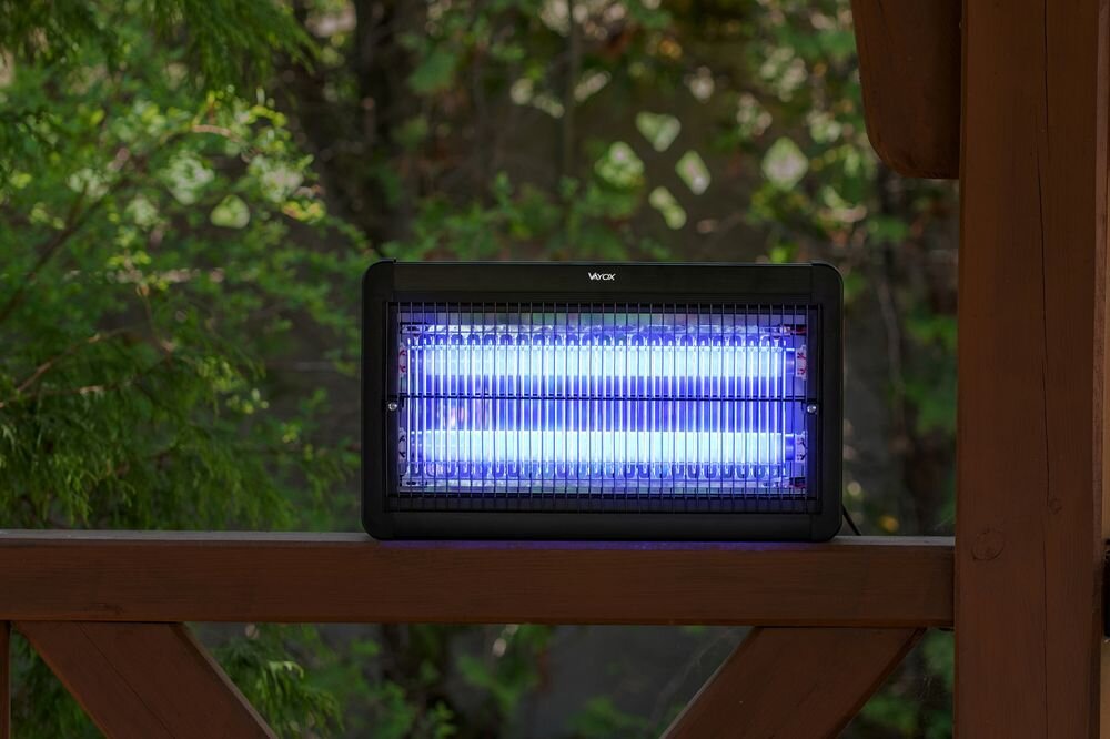 Lampa owadobójcza z niebieskim światłem umieszczona na drewnianej powierzchni. W tle widoczne są zielone liście i drewniana konstrukcja.