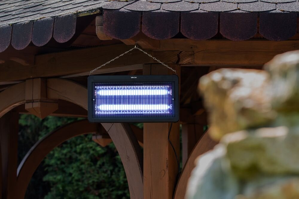 Lampa owadobójcza wisi na łańcuchu pod drewnianą konstrukcją, emitując niebieskie światło. Na obudowie widoczne jest logo \'VAYOX\'.