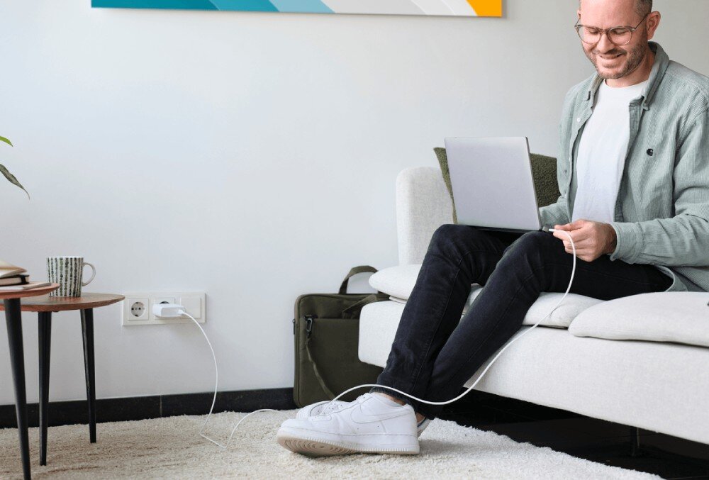 Zasilacz do laptopa TRUST Maxo 25140 100W Mężczyzna w okularach pracujący na kanapie z laptopem podłączonym kablem do ładowarki wpiętej w dolne gniazdko ścienne