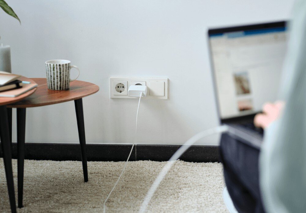Zasilacz do laptopa TRUST Maxo 25140 100W Kadr przedstawiający stolik kawowy oraz ładowarkę wpiętą do kontaktu i połączoną kablem z laptopem osoby siedzącej na dywanie