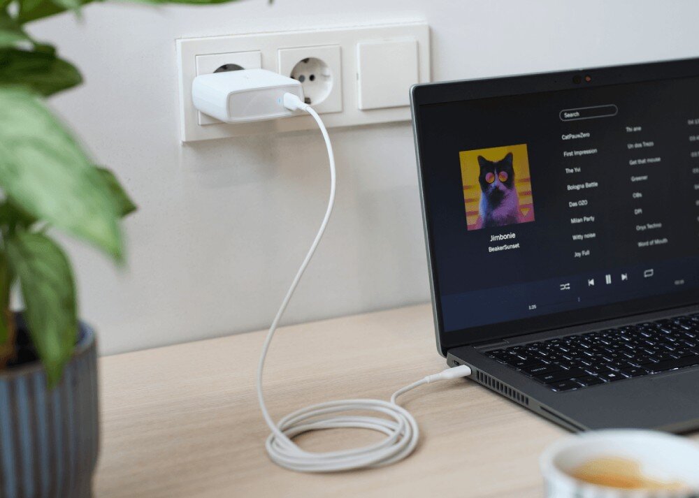 Zasilacz do laptopa TRUST Maxo 25140 100W Biała ładowarka sieciowa w gniazdku elektrycznym zasilająca laptopa z otwartym odtwarzaczem muzyki stojącego na biurku obok rośliny