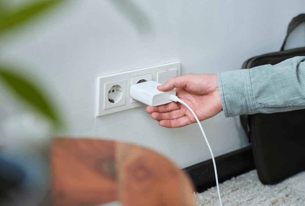 Zasilacz do laptopa TRUST Maxo 25140 100W Zbliżenie na dłoń wkładającą białą ładowarkę do ściennego gniazdka elektrycznego