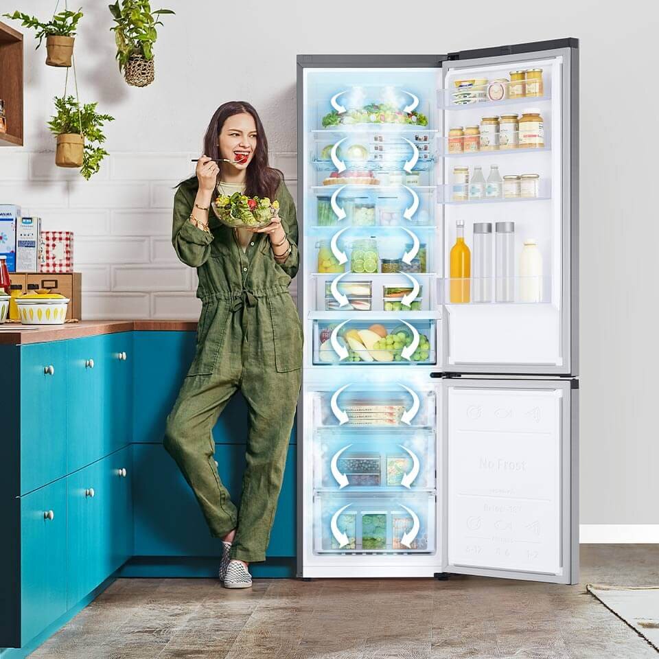 Kobieta stoi obok otwartej lodówki w kuchni, trzymając miskę sałatki. Wnętrze lodówki jest wypełnione różnymi produktami spożywczymi, a na drzwiach widoczne są butelki i słoiki.