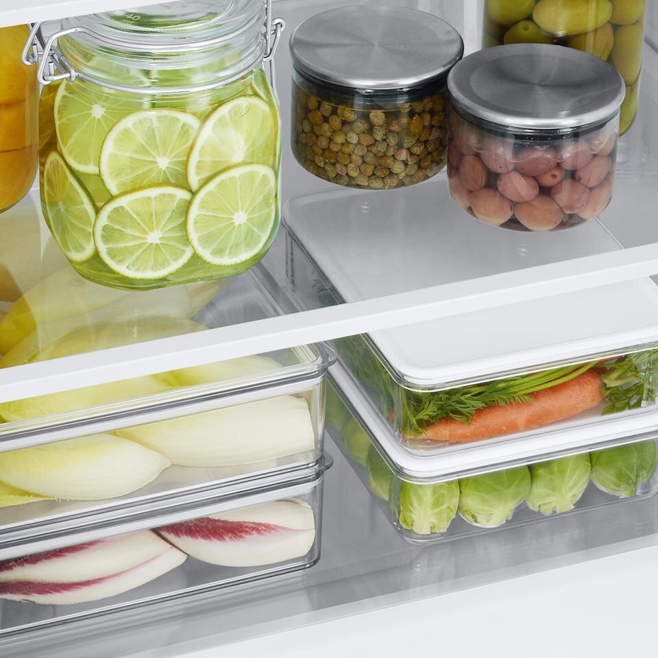 W lodówce znajdują się słoiki z pokrojonymi limonkami, oliwkami i innymi produktami spożywczymi oraz pojemniki z warzywami, takimi jak marchewki i brukselka. Wszystko jest uporządkowane na półkach.