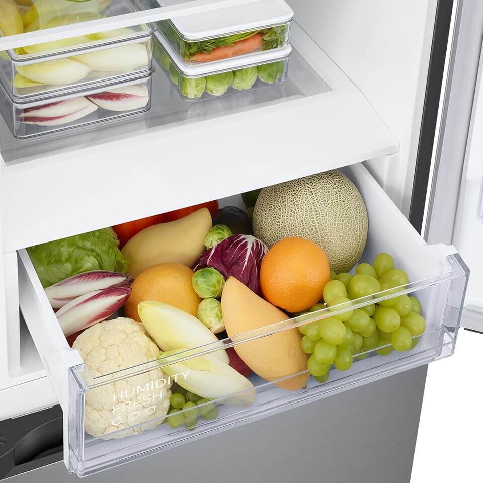 Otwarta szuflada lodówki wypełniona różnymi owocami i warzywami, takimi jak winogrona, pomarańcze, melon i sałata. Na szufladzie widoczny jest napis \'HUMIDITY FRESH\'.