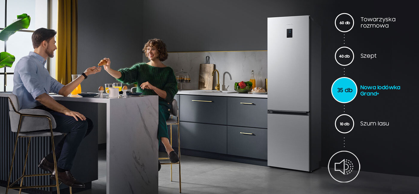 W kuchni przy stole siedzą dwie osoby, a obok znajduje się lodówka w metalicznym grafitowym kolorze. Obok lodówki widoczna jest grafika z poziomami hałasu: 60 dB Towarzyska rozmowa, 40 dB Szept, 35 dB Nowa lodówka Grand+, 10 dB Szum lasu.