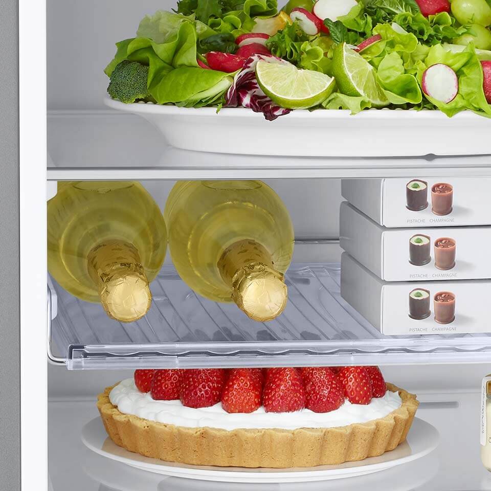 Wnętrze lodówki z widocznymi produktami spożywczymi: na górnej półce znajduje się talerz z sałatką, poniżej dwie butelki wina oraz pudełka z lodami, a na dolnej półce tarta z truskawkami.
