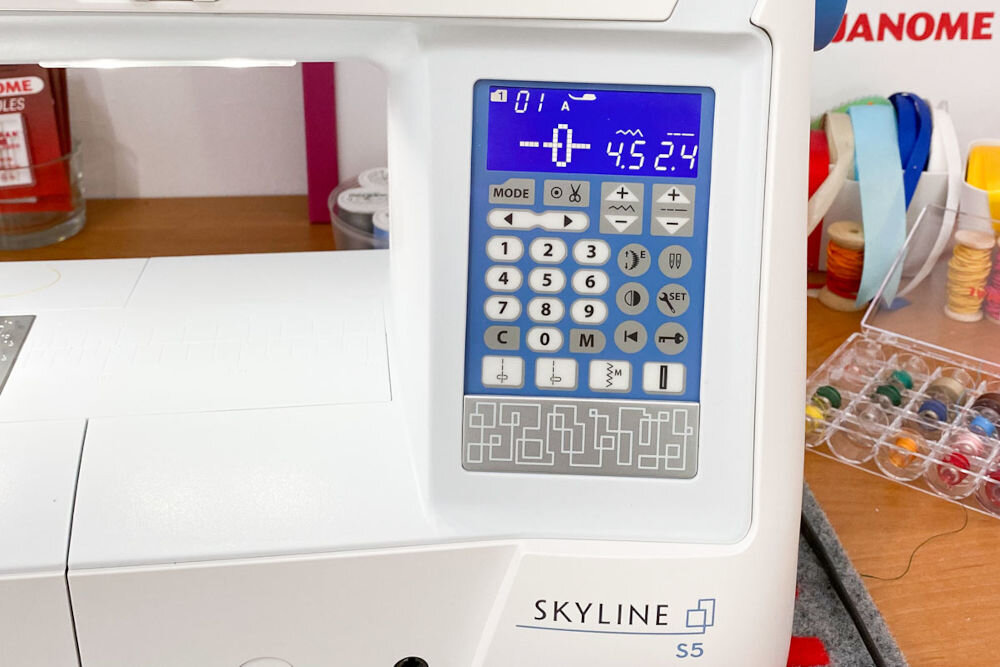 Maszyna do szycia JANOME Skyline S5 ekran niebieskie elementy przyciski cyfry znaki podświetlany ekran LCD wybrany ścieg jego parametry rekomendacje dotyczące użycia odpowiedniej stopki standardowe wyposażenie maszyny czytelny wyświetlacz łatwa intuicyjna obsługa szybkie ustawienie parametrów szycia