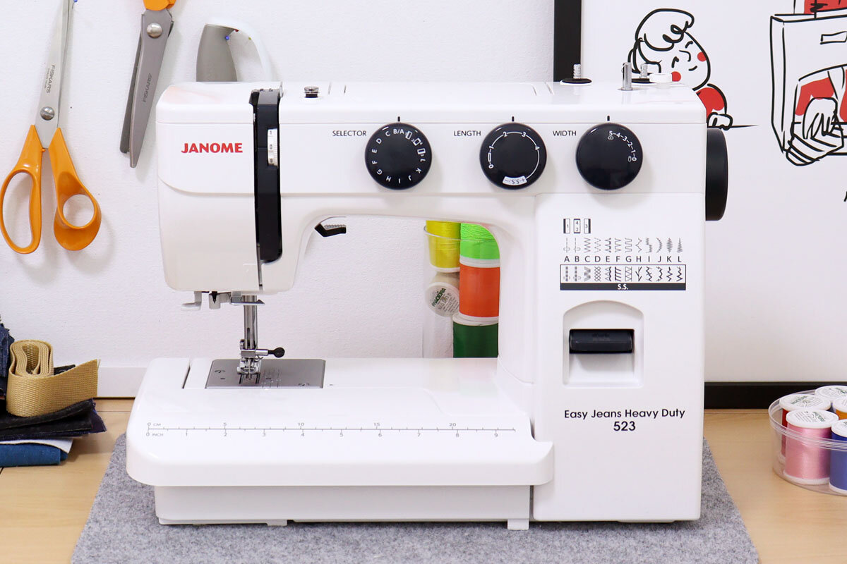 Na obrazie znajduje się maszyna do szycia z napisem \'JANOME\' oraz \'Easy Jeans Heavy Duty 523\'. W tle widoczne są nożyczki, nici i inne akcesoria krawieckie.