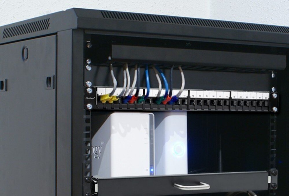 Patch panel QOLTEC 54516 Widok wnętrza metalowej szafy RACK z zamontowanym panelem sieciowym, do którego podłączone są kolorowe przewody RJ45 oraz widoczne poniżej urządzenia sieciowe z podświetleniem diod, opis