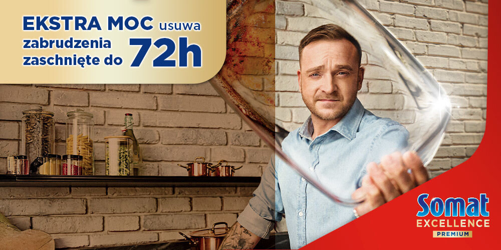Tabletki do zmywarek SOMAT Excellence Premium 5w1 - 42 szt. Ujęcie mężczyzny w niebieskiej koszuli (prawdopodobnie ambasadora marki) trzymającego lśniącą, czystą szklankę. W tle kuchnia ze ścianą z białej cegły. W lewym górnym rogu hasło: EKSTRA MOC usuwa zabrudzenia zaschnięte do 72h. Logotyp Somat w prawym dolnym rogu